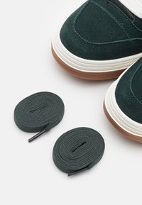 Zapatillas de ante verde con suelas de goma blanca. Dos cordones verdes enrollados con puntas negras situados al lado de los zapatos. Presentan detalles perforados.