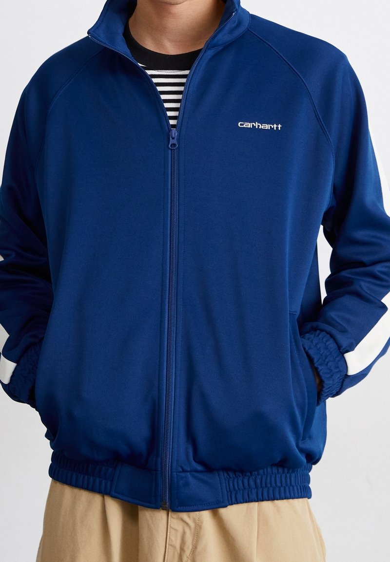 Mies, joka käyttää sinistä vetoketjullista takkia, jossa on valkoisia raitoja hihoissa, kädet taskuissa, mustavalkoisen raidallisen paidan ja vaaleat housut päällään.