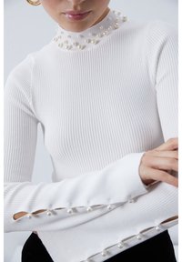 Biały prążkowany top z długim rękawem, z wysokim dekoltem ozdobionym perłami oraz wycięciami na rękawach z perłowymi detalami na mankietach.