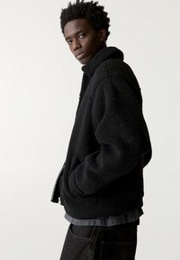 Veste en polaire noire avec un col montant, une fermeture éclair à l'avant et des poches latérales. Présente une texture douce et une coupe décontractée, portée sur une chemise grise.