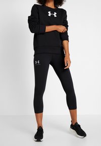 Sudadera negra con un logo blanco, combinada con leggings capri negros que tienen un pequeño logo. Ambos están hechos de una tela suave y tienen un diseño ajustado.