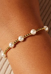 Gouden armband met parels, afwisselend ronde parels en gouden kralen, weergegeven tegen een gebruinde huid, met een gladde, glanzende afwerking.