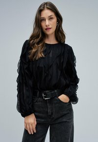 Blusa nera, lavorata con dettagli a rouches e maniche a palloncino, abbinata a jeans in denim scuro e una cintura nera con una fibbia metallica.