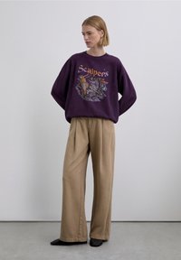 Lilla oversized sweatshirt med grafisk print, der viser en fugl og tekst; kombineret med beige bukser med vide ben og sorte slip-on sko.