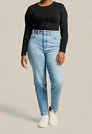 Persona con vaqueros de tiro alto azul claro, camiseta negra de manga larga y zapatillas blancas de pie frente a un fondo liso.