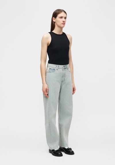 Γυναίκα που στέκεται φορώντας μαύρο αμάνικο τοπ, ανοιχτό μπλε τζιν με φαρδιά πόδια και μαύρα μοκασίνια σε ουδέτερο ανοιχτό φόντο.