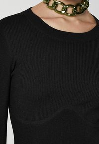 Maglietta nera a maniche lunghe con coste, caratterizzata da un colletto rotondo e dettagli di design curvi. Accompagnata da una collana di catena spessa verde.