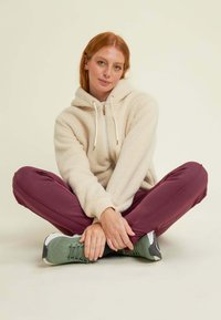 Felpa in pile beige con cappuccio, cordini e zip, abbinata a pantaloni elasticizzati bordeaux e scarpe sportive verdi con accenti bianchi.