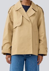 Beige oversized jakke med klippet krave, frontlåg og lommer; har skulderepauletter og løs pasform; parret med blå denim.