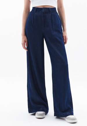 Donkerblauwe wijde spijkerbroek van denim, met een hoge taille, plooien aan de voorkant en een enkele knoopsluiting. Draagt grijze sneakers.