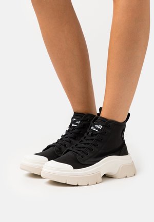 Baskets montantes noires avec une semelle blanche épaisse, un dessus en toile mate et des lacets noirs. Présente une étiquette logo sur la languette.