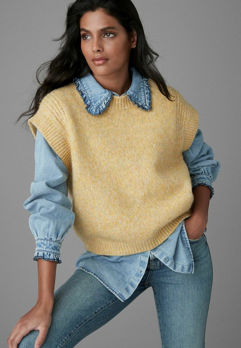 Un pull en maille jaune porté sur une chemise en denim bleu clair avec un col volanté, associé à un jean en denim ajusté, le tout sur un fond gris.