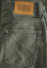 Buzunar spate al blugilor din denim verde cu o etichetă din piele maro pe care scrie "JACK & JONES" cusută deasupra buzunarului.