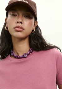 Young woman wearing a brown cap with "PROCESS," gold hoop earrings, a chunky purple stone necklace, and a pink crew neck t-shirt.