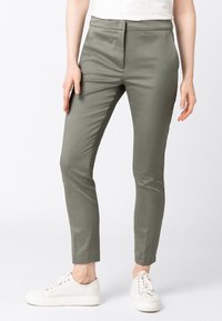 Pantaloni aderenti verdi oliva in tessuto liscio, con vita piatta, tasche laterali e taglio alla caviglia, abbinati a sneakers bianche.