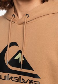 Felpa color marrone chiaro con coulisse e logo nero con un design di montagna. Texture liscia con inserti metallici sulla coulisse.