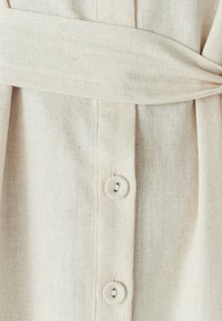 Béžové ľanové šaty s opaskom, obsahujúce dva malé okrúhle gombíky a jemnú textúru, navrhnuté s plisovaním pre zvýraznenie detailov.