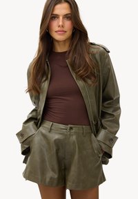 Femme portant une veste en cuir vert olive et un short avec un haut marron ajusté, debout les mains dans les poches contre un fond uni.