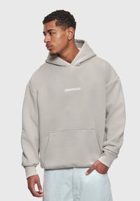 Joven con cabello corto y rizado que lleva una sudadera con capucha beige "DROPSIZE" y pantalones de color claro, mirando hacia la izquierda con una mano en el bolsillo.