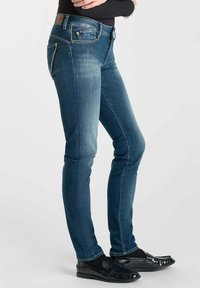 Persoon in aansluitende blauwe jeans, zwart longsleeve shirt en zwarte instapschoenen, staande tegen een effen lichte achtergrond.