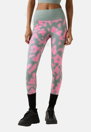 Frau trägt graue Leggings mit hohem Bund und leuchtend pinkem abstraktem Muster sowie schwarze Sportschuhe, steht vor weißem Hintergrund.