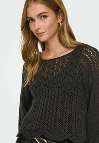 Pull en tricot noir avec un col rond, des manches longues et des motifs ajourés, présentant un ourlet en forme de vagues et une finition texturée.