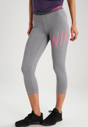 Kvinde iført grå korte leggings med pink „NIKE“-tekst på låret og sorte sportssko, stående i afslappet stilling.