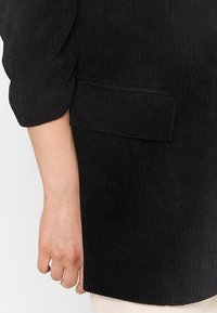 Gros plan sur une manche de veste noire côtelée et une poche à rabat, montrant une main nue reposant à côté.