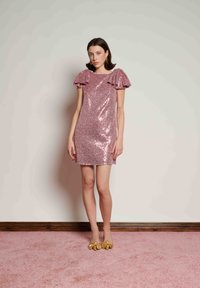 Φόρεμα με παγιέτες σε ροζ χρώμα που λάμπει, με φουσκωτά μανίκια, εφαρμοστή γραμμή και τελείωμα πάνω από το γόνατο. Φοριέται με κίτρινα παπούτσια με φιόγκο.