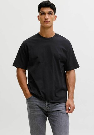 Junger Mann, der nach vorne blickt und ein schlichtes, schwarzes, übergroßes T-Shirt sowie graue Jeans trägt, mit einer Hand in der Tasche.