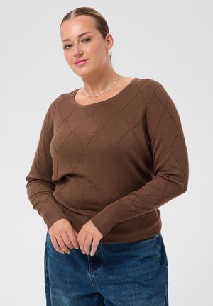 Femme portant un pull marron à manches longues avec un motif en losanges et un jean bleu, debout, mains jointes, sur un fond gris uni.