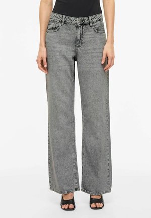 Person iført højtaljede, vidde grå acid-wash jeans med frontlommer og sorte åbne hæle, stående mod en hvid baggrund.