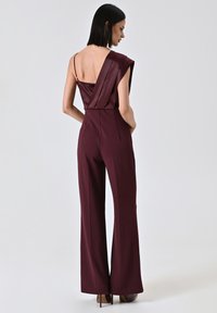 Combinaison bourgogne avec une texture satinée, présentant un design à dos drapé et des jambes larges. Les bretelles sont fines et il y a des coutures sur les côtés.