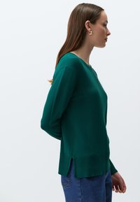 Zielony sweter z dzianiny, z długimi rękawami, okrągłym dekoltem, bocznymi rozcięciami u dołu i prążkowanymi akcentami. Połączony z niebieskimi dżinsami.