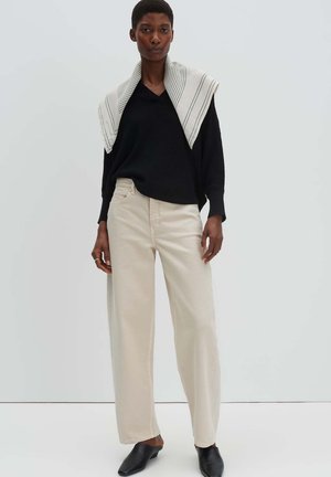 Persona alta che indossa un maglione nero a scollo a V, pantaloni larghi color crema, scarpe slip-on nere e una sciarpa bianca a righe drappeggiata sulle spalle.