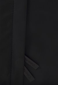 Il tessuto nero presenta una texture liscia con cuciture visibili. Due etichette con testo in rilievo sono parzialmente visibili sul bordo.