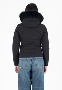 Chaqueta acolchada negra con capucha, con un ribete de piel sintética. El diseño es ajustado en la cintura, con jeans anchos azules visibles en la parte inferior.