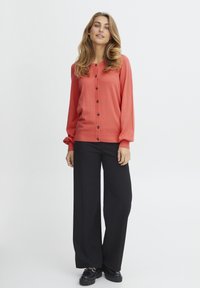 Gilet coral à boutons avec des manches longues, des poignets côtelés et une rangée de boutons foncés, assorti à un pantalon large noir et des chaussures à plateforme.