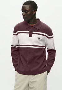 En ung mand iført en rød og hvidstribet langærmet polo sweater og beige bukser, som kigger til venstre med hænderne i lommerne.
