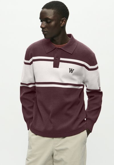 Jeune homme portant un pull polo rayé bordeaux et blanc à manches longues et un pantalon beige, regardant à sa gauche les mains dans les poches.