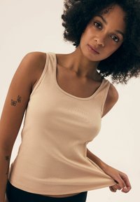 Beżowy top na ramiączkach z okrągłym dekoltem i dopasowanym krojem, z gładką fakturą, noszony włożony w talię, podkreślający sylwetkę.