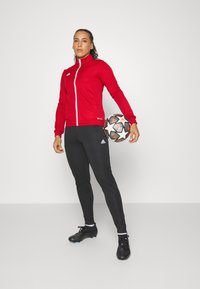 Rød atletisk jakke med fuld lynlås, sorte tætsiddende bukser og sorte fodboldstøvler. Spilleren holder en mønstrede fodbold dekoreret med stjerner.