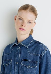 Tumši zilas džinsa krekls ar norādītu apkakli, divām krūšu kabatām un šuvumu detaļām. Virs kreisās kabatas atrodas logotipa marķējums.