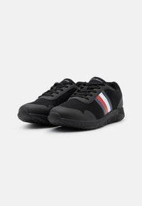Chaussures de sport noires avec panneaux en mesh, bout rond et semelles en caoutchouc. Présentent des bandes blanches, bleues marine et rouges sur les côtés.