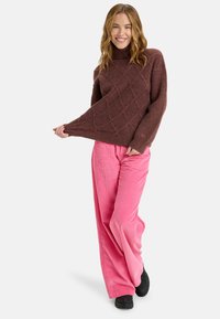 Suéter marrón de punto trenzado con cuello alto, combinado con pantalones de pana anchos rosas. Modelo vistiendo calzado negro y sonriendo.