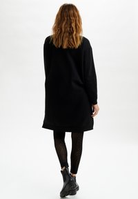 Czarna oversize sukienka-sweterek z długimi rękawami, miękkimi mankietami i okrągłym dołem, zestawiona z czarnymi legginsami o teksturowanej powierzchni oraz botkami za kostkę.