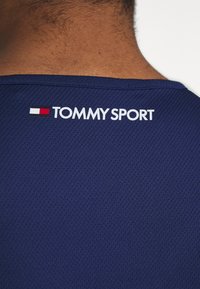 Nuca e spalle con una maglietta sportiva blu navy con il testo bianco "TOMMY SPORT" e un logo rosso, bianco e nero.
