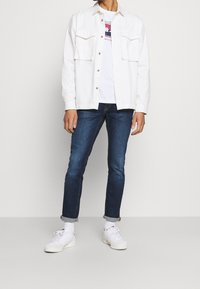 Chaqueta de denim blanca con dos bolsillos en el pecho, llevada sobre una camiseta gráfica blanca. Unos jeans oscurecidos de color azul y zapatillas blancas completan el atuendo.