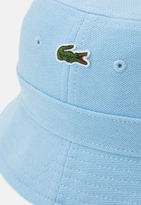Lyseblå bøttehatt laget av strukturert stoff, med en liten grønn krokodille-logo og to ventilasjonshull på siden.