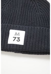 Tumedast merest kootud müts, millel on tekstuuriline muster. Kaasas valge silt, millel on mustalt trükitud "M 73". Lihtne ümmargune kuju.